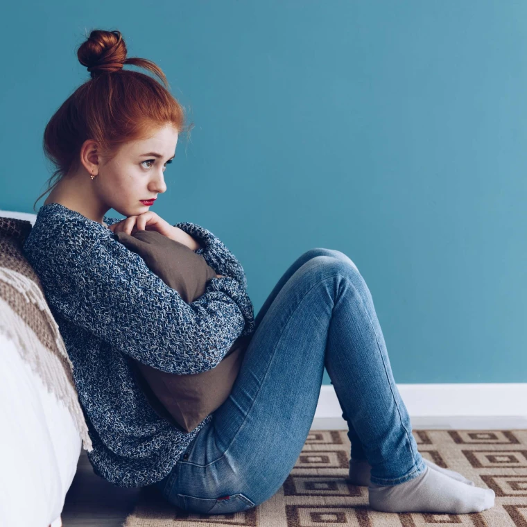 Woman sat on the floor, leaning against her bed and clutching a pillow